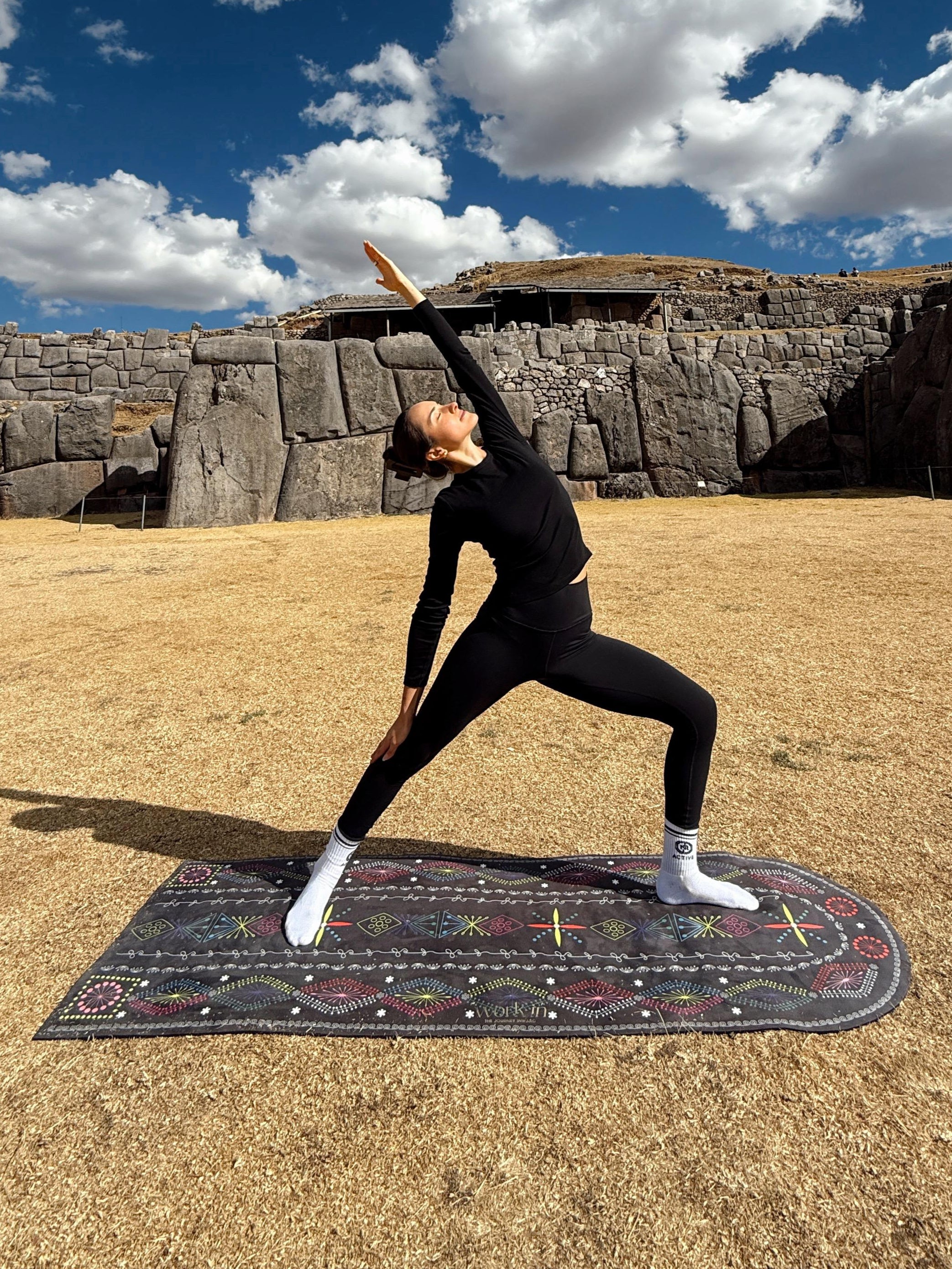 Person practicing yoga in front of ancient stone walls under a blue sky with clouds.